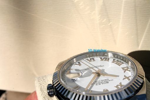 IMG_6716 | SD Watches Close-up of a silver luxury watch with Roman numerals and a ridged bezel, seen at an angle on a beige background (watch face visible).