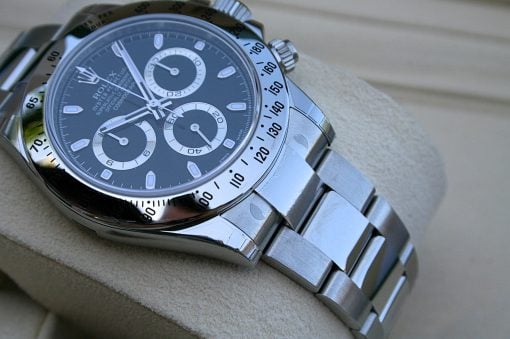IMG_6823 | SD Watches Close-up of a silver Rolex chronograph wristwatch with a black dial, white subdials, and a tachymeter bezel resting on a beige surface fields.