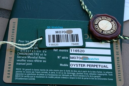 IMG_6856 | SD Watches Close-up of a Rolex warranty card with a barcode, serial number, and a red wax seal attached by a cord, on a green surface.