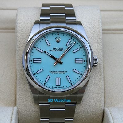 Rolex Oyster Perpetual watch with a turquoise dial on a beige display cushion, stainless steel bracelet visible.