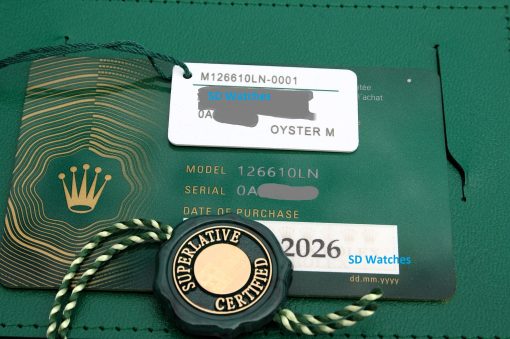IMG_6918 | SD Watches White tag with model number M126610LN-0001 placed on a green Rolex warranty card; blue 'SD Watches' text obscures part of the tag.