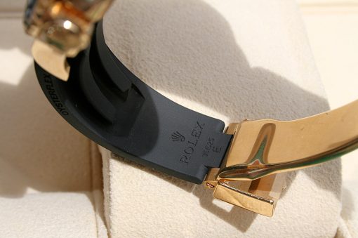IMG_6936 | SD Watches Close-up of a black rubber watch strap with a gold-tone deployment clasp, beige display surface, ROLEX partially visible on the strap.
