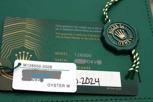 IMG_7115 | SD Watches Close-up of a Rolex purchase card with a green background, gold line pattern, and a green seal tag attached by a braided string with a white label partially overlaid.