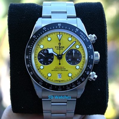 Tudor Genève chronograph with a bright yellow dial, black subdials, and a stainless steel bracelet on a black display cushion.