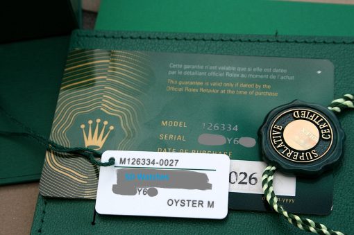 IMG_7293 | SD Watches Close-up of a green Rolex warranty card with gold text, white tag showing model M126334-0027, and a black circular seal reading 'SUPERLATIVE' or 'SUPERLATIVE OFFICIALLY CERTIFIED'
