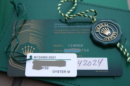 IMG_7439 | SD Watches Close-up of a blue Rolex tag with crown logo attached to a green card, braided string visible, with model/serial numbers faintly showing beneath.