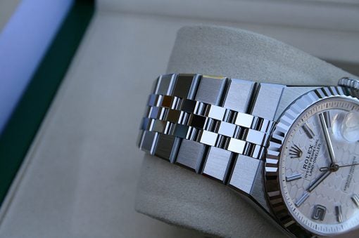 IMG_7490 | SD Watches Close-up of a silver Rolex-style wristwatch with a metal link bracelet resting on a cushioned display surface, showing the textured bezel and dial details behind a glass face.