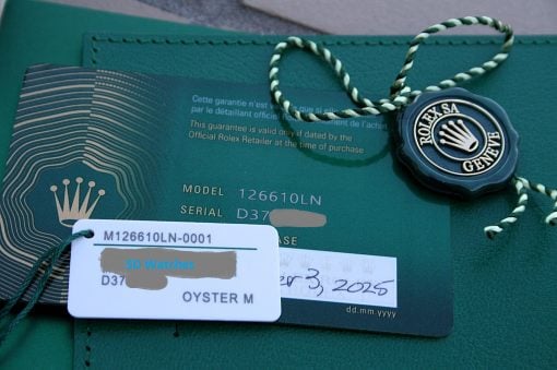 IMG_7513 | SD Watches Close-up of a green Rolex warranty card with a black seal attached by a green-and-white braided cord, showing model and serial numbers and a white tag partly obscured.